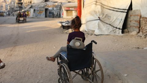 En flicka i rullstol på en gata i Gaza, med skadade byggnader i bakgrunden.