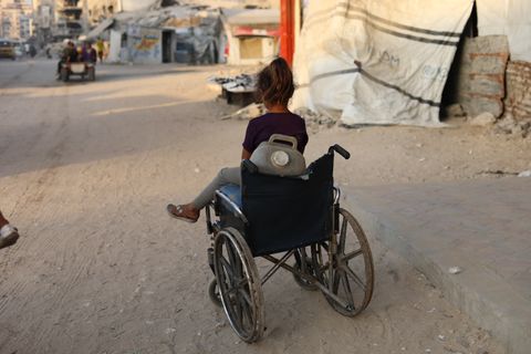 En flicka i rullstol på en gata i Gaza, med skadade byggnader i bakgrunden.