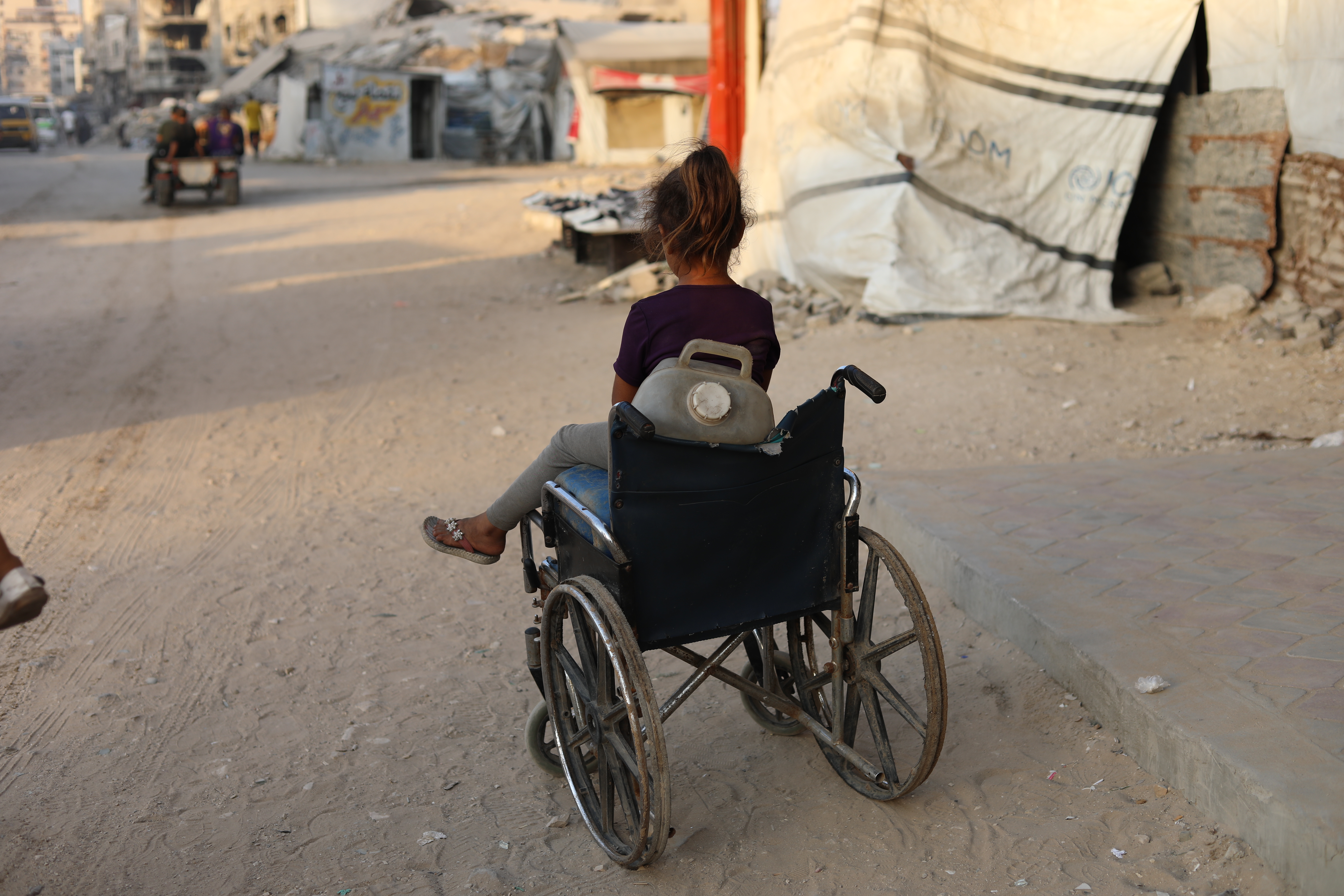 En flicka i rullstol på en gata i Gaza, med skadade byggnader i bakgrunden.