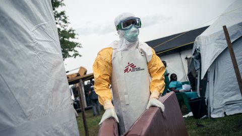 En person iklädd skyddsutrustning med Läkare Utan Gränsers logga på, står framför isoleringstält och håller i två madrasser.