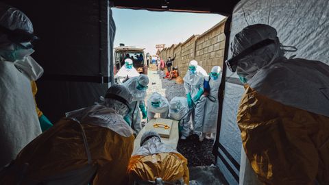 Medarbetare i skyddsutrustning från Läkare Utan Gränser bär ut en kista i staden Bunia, Kongo-Kinshasa, med en person som har avlidit till följd av ebola. Utanför dörren tas kistan emot av annan personal.