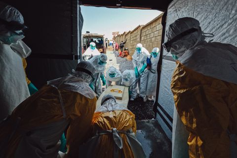 Medarbetare i skyddsutrustning från Läkare Utan Gränser bär ut en kista i staden Bunia, Kongo-Kinshasa, med en person som har avlidit till följd av ebola. Utanför dörren tas kistan emot av annan personal.