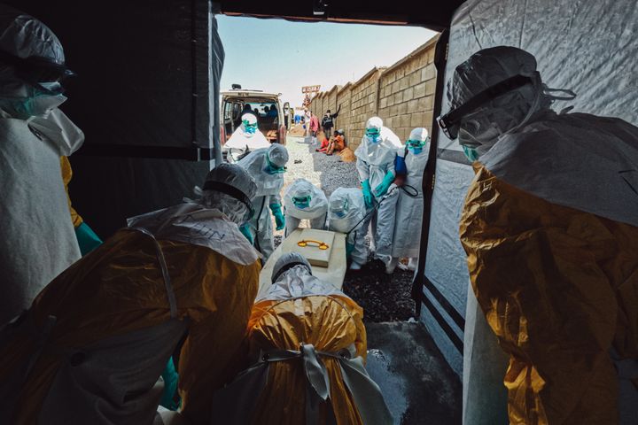 Medarbetare i skyddsutrustning från Läkare Utan Gränser bär ut en kista i staden Bunia, Kongo-Kinshasa, med en person som har avlidit till följd av ebola. Utanför dörren tas kistan emot av annan personal.