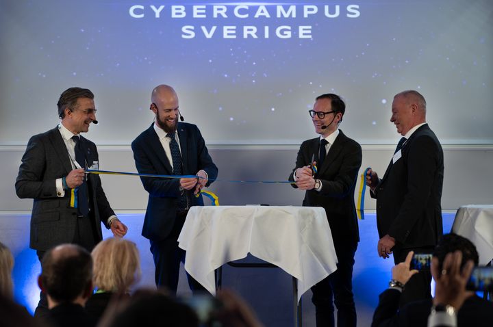 Four men dressed in suits cut a blue and gold ribbon on stage.