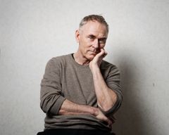 En man med kort hår och grå tröja sitter med handen vid hakan och tittar mot kameran.