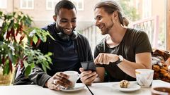 Två personer sitter vid ett bord på ett kafé, leende och tittar på en smartphone.