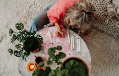 En person sitter på golvet och ritar på ett papper vid ett runt soffbord, omgiven av gröna växter och tända ljus.