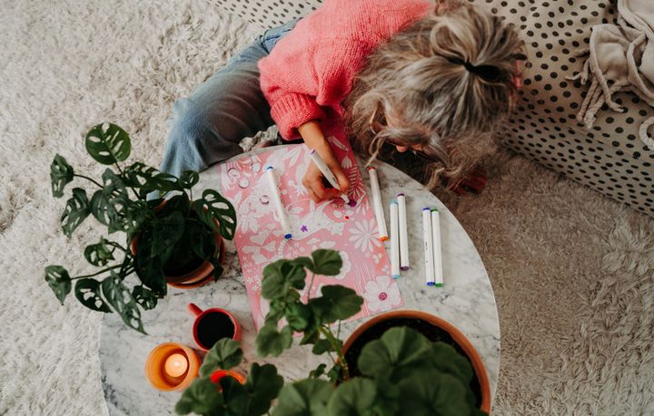 En person sitter på golvet och ritar på ett papper vid ett runt soffbord, omgiven av gröna växter och tända ljus.