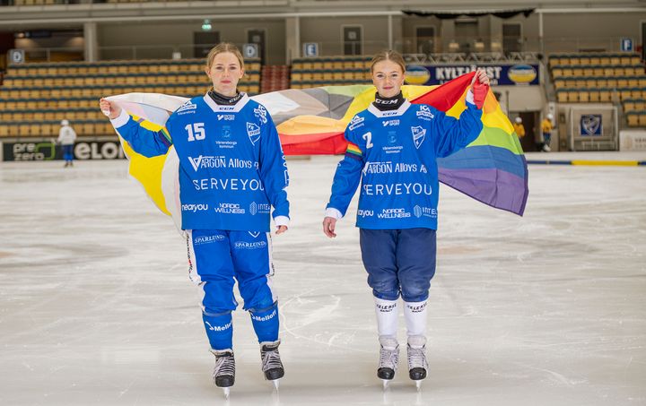 Två unga tjejer i IFK Vänersborgs klubbkläder åker skridskor med en hbtqi-flagga bakom sig.