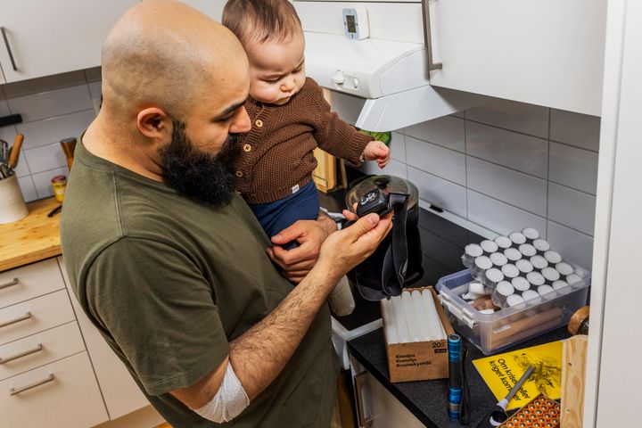En man står i ett kök med en bebis på armen. På bänken finns saker för att hantera kriser.