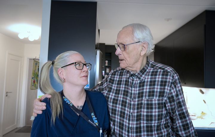 En kvinnlig hemtjänstpersonal står intill en äldre man. De ser på varandra.