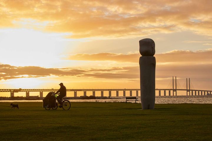 Cyklist och hund vid stranden med en stor staty och en bro i solnedgången.