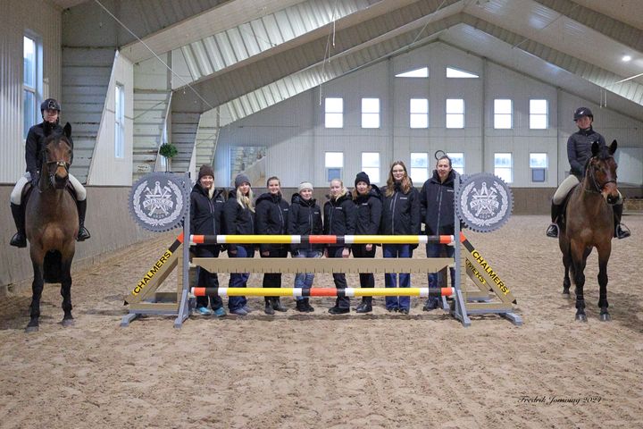 Gruppfoto i ett ridhus med åtta personer stående bakom ett hinder och två ryttare på hästar.