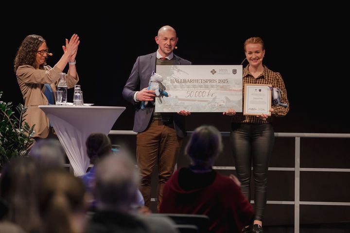 En man och en kvinna från företaget Ekolution står på scenen under Hästföretagarforum och håller i en stor check och ett diplom vid prisutdelningen. Bredvid applåderar en kvinnlig moderator.