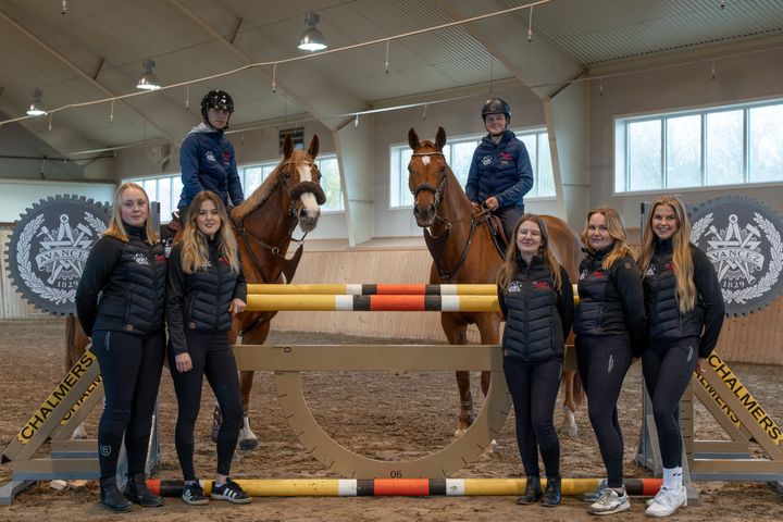 En grupp människor i ett ridhus, två sitter på hästar och andra står bredvid ett hinder.