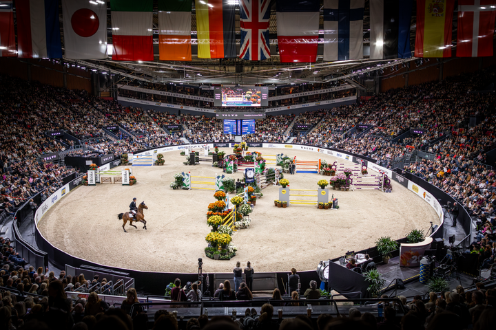 En ryttare hoppar hinder på en häst i en fullsatt inomhusarena med publiken och flera nationella flaggor i taket.