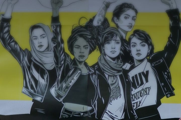 Bild på fem kvinnor som står tätt tillsammans och höjer sina nävar i en kraftfull gest av motstånd och solidaritet. Bilden är svar och vit med en gul bakgrund.