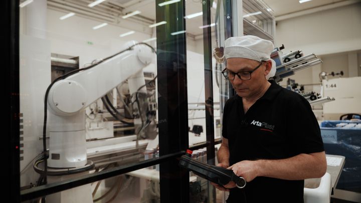 En man i arbetskläder och mössa står framför en glasruta och använder en surfplatta nära en industrirobot inuti en fabrik.