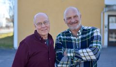 Två leende män framför en gul vägg.