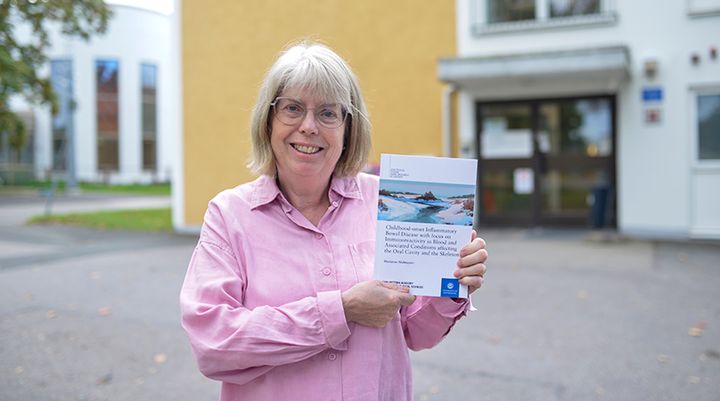 Kvinna står utanför en byggnad, håller upp en bok och ler