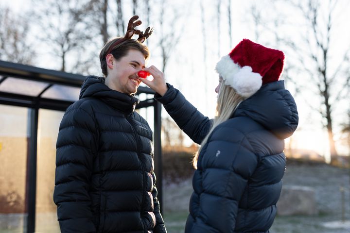 Två personer i vinterkläder. Den ena personen har renhorn och en röd näsa, medan den andra sätter på en tomteluva.