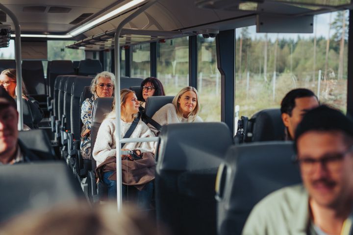 En grupp människor sitter på en buss, med skog utanför fönstren. Några av dem ser glada ut och pratar med varandra.