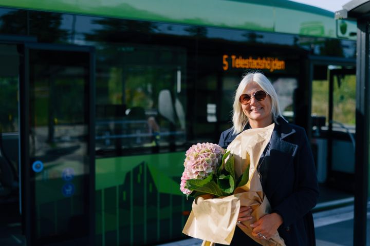 Kvinna med solglasögon håller en bukett blommor framför en grön stadsbuss.