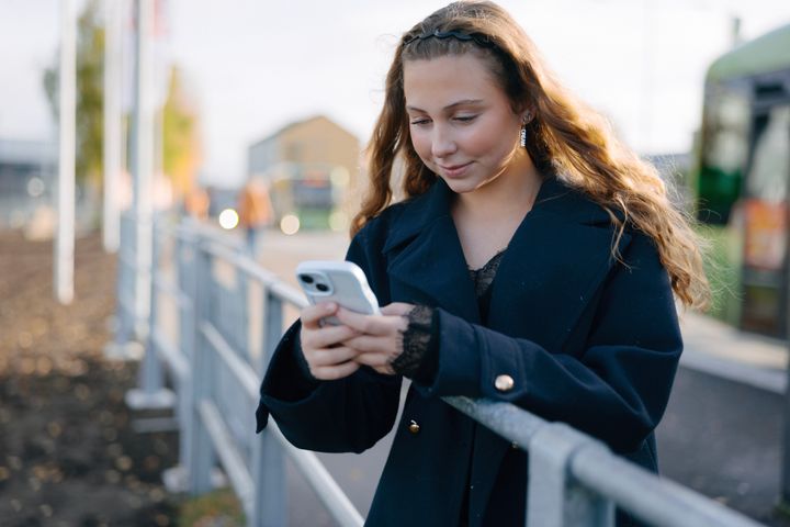 En ung kvinna står vid ett staket och tittar på sin mobiltelefon, med en buss i bakgrunden.