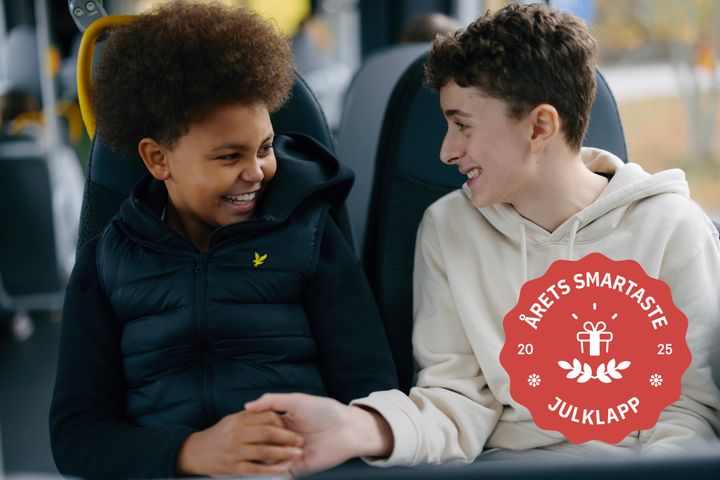 Två barn sitter och ler på en buss. En röd julklappsetikett bredvid dem säger "Årets smartaste julklapp 2025".