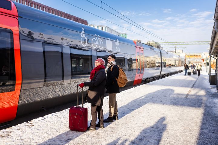 Två personer står på en snötäckt perrong med resväskor, bredvid ett stillastående tåg.
