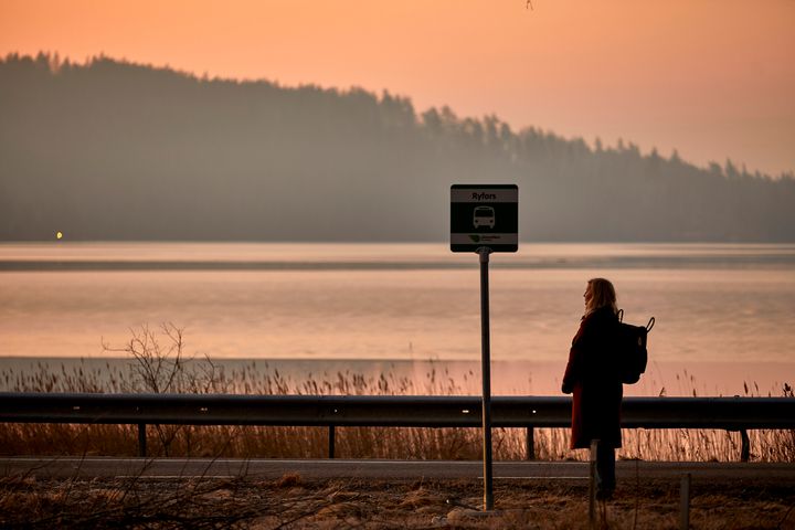 Kvinna väntar vid en busshållplats vid solnedgång med en sjö i bakgrunden.