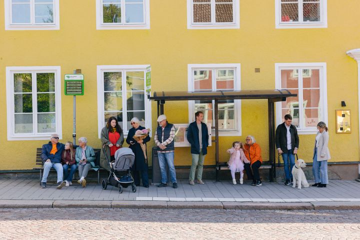 En grupp människor väntar vid en busskur framför en gul byggnad.