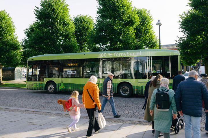 En grön buss märkt "100 % förnybar el" står vid en hållplats. Flera människor går på trottoaren bredvid bussen.