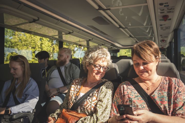En grupp människor sitter på rad på en buss, tittar på sina mobiltelefoner och samtalar. Det är soligt ute.