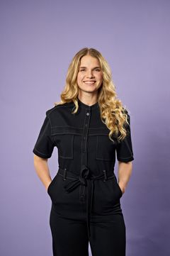 Kvinna i mörk jumpsuit står mot en lila bakgrund.