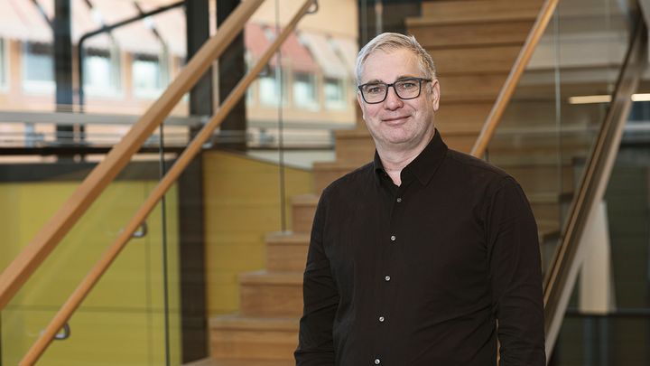 Man i svart skjorta och glasögon framför en trappa.