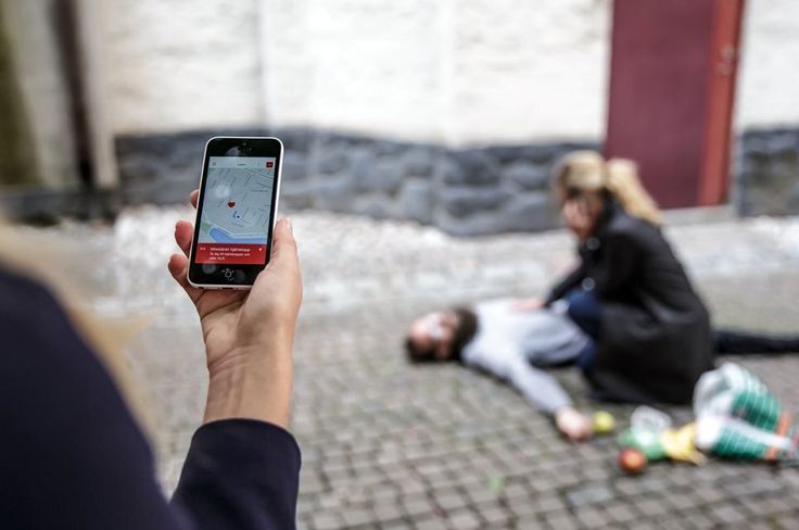 En mobiltelefon med appen sms-livräddare i bild, där en man skymtas i bakgrunden som fallit ihop i plötsligt hjärtstopp där kvinna gör känner efter puls och ringer ambulans.