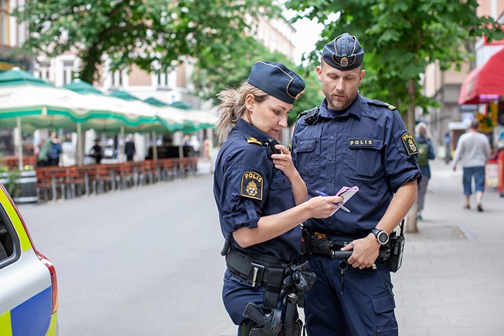 Två poliser på ett torg som kollar i ett block och lyssnar på en polisradio.