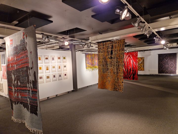 Utställningsrum på Hallstahammars konsthall med hängande textilkonstverk och inramade bilder på väggarna.