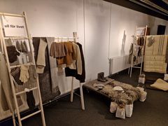 Bild av utställningen "Ull för livet" med olika klädesplagg och textilier gjorda av ull, upphängda och utställda i en konsthall.