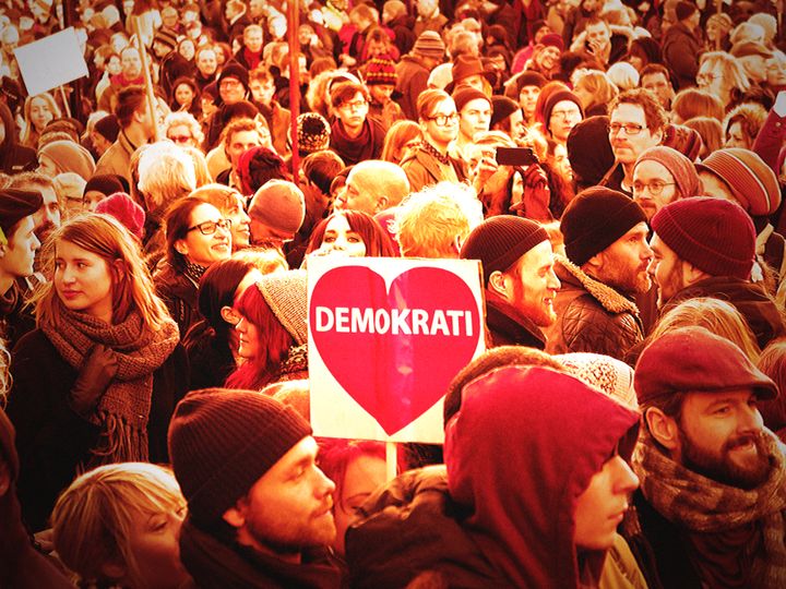 En folkmassa demonstrerar, en person håller en skylt med ett rött hjärta och texten "Demokrati".