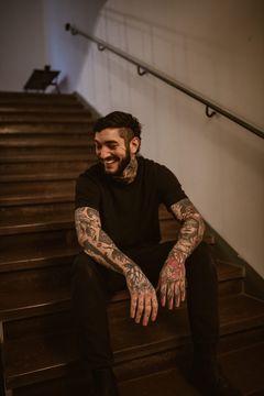 Attila Yoldas sitter leende i en trappa, hans armar är täckta av tatueringar
