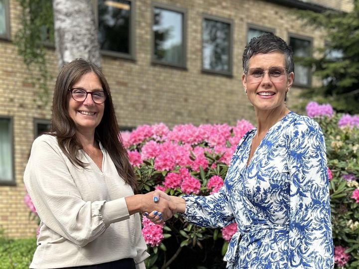 Två kvinnor skakar hand framför blommande buskar och en byggnad.