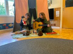 Två kvinnor sitter på golvet i ett lekrum; en spelar gitarr medan den andra håller en docka.