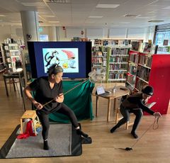 Två personer i svarta kläder uppträder med en dockteater i ett bibliotek.