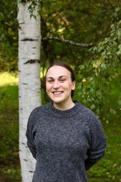 En person står leende framför en björk, med grönska i bakgrunden.