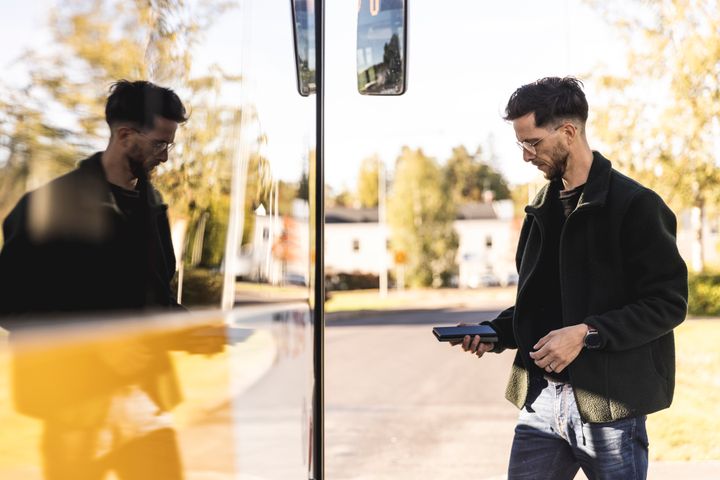 En man är på väg att gå på en buss, han håller sin telefon i handen. I bussens fönster blir det en spegling av honom.