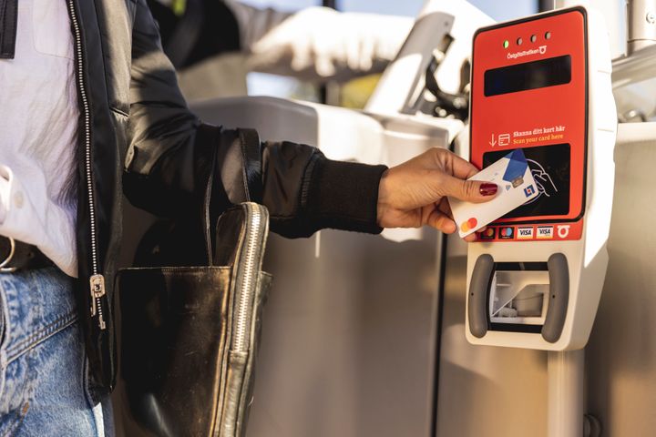 En arm som håller fram ett betalkort mot en biljettläsare ombord på en buss.