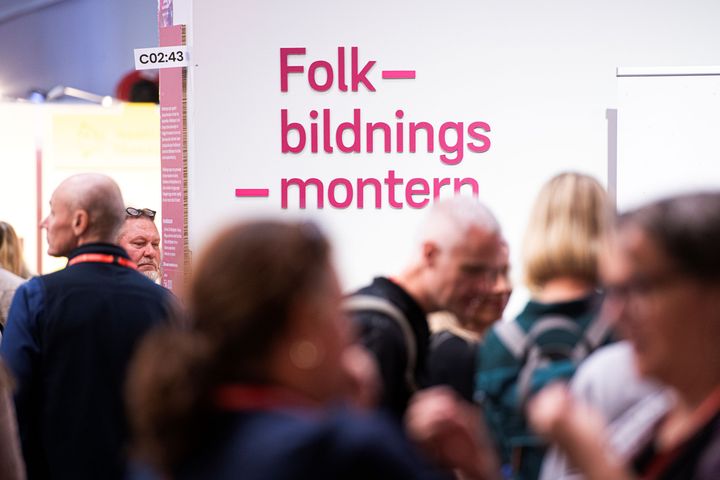 En bild på Folkbildningsmontern från förra årets bokmässa: "Folkbildningsmontern" i lila text på på grå vägg. Människor i förgrunden.