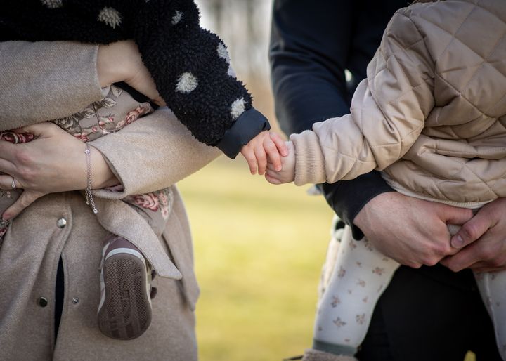 Två små barn håller varandra i handen i vuxnas famn.
